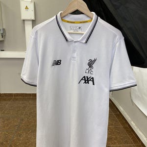 May include: A white short-sleeved polo shirt featuring a black New Balance logo on the right chest and a black Liverpool Football Club crest with the text L.F.C. and AXA below it on the left chest. The collar and sleeve cuffs have black trim.