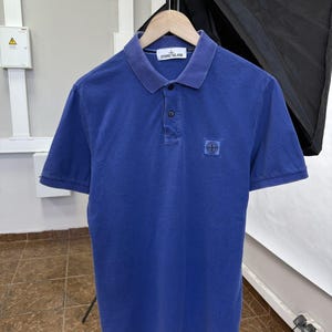 May include: A blue short-sleeved polo shirt featuring a classic collar, a two-button placket, and a Stone Island brand patch on the left chest. The garment includes a white interior neck label with the Stone Island logo and text in black.