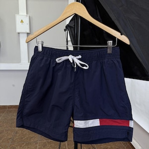 May include: Navy blue swim shorts with a white drawstring and a white, red, and navy blue stripe detail on the leg. The shorts are hanging on a wooden hanger.