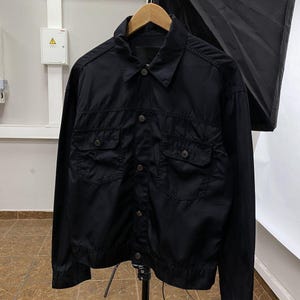 May include: Black jacket with two chest pockets and button closures. The jacket is hanging on a wooden hanger. The jacket has a collared neckline and long sleeves. The jacket is made of a lightweight material.