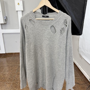 May include: A light grey sweater with distressed detailing, including holes at the chest and along the sleeves. The sweater is displayed on a wooden hanger. The sweater has a ribbed hem and cuffs.
