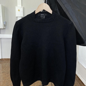 Può includere: Un maglione nero girocollo con polsini e orlo a coste, appeso a una gruccia di legno. Il maglione ha un design semplice e classico. Il marchio "GUCCI" è visibile sull'etichetta.