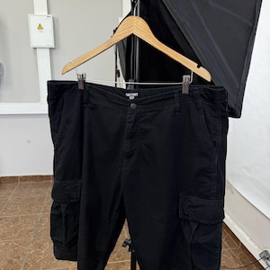 May include: Black cargo shorts featuring a button closure, two side pockets with flaps, and two front pockets. The interior waistband label displays the text Carhartt WIP. The shorts are suspended from a wooden hanger by metal clips.