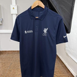 May include: A dark navy blue short-sleeved polo shirt featuring a white Nike swoosh on the left sleeve, a white Standard Chartered logo on the right chest, and a white Liverpool Football Club L.F.C. crest on the left chest with a collar.