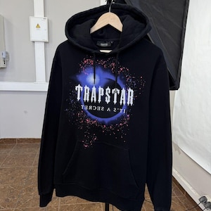 May include: A black hooded sweatshirt featuring a graphic print on the chest with the word TRAPSTAR in white gothic lettering above the text IT'S A SECRET. The design includes a blue circular nebula effect with pink and blue paint splatter accents.