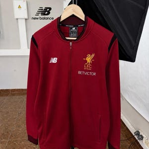 May include: A dark red zip-up track jacket featuring a ribbed collar, black shoulder accents, and long sleeves. The chest displays a white New Balance logo and a gold Liverpool FC 125 Years crest with the text BETVICTOR printed below in white.