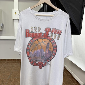 May include: A white short-sleeve t-shirt featuring a graphic print with the text Repeat 3-Peat and NBA Champions. The design includes a basketball, a city skyline, and a red bull head logo, along with six small trophy icons above the text.