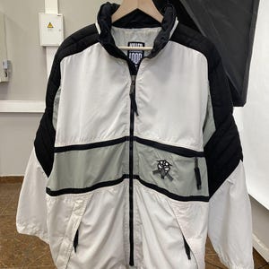 May include: A white, black, and grey jacket with the text "KILLER LOOP". The jacket features a full zip, a high collar, and a basketball graphic. It has two front pockets and is hanging on a wooden hanger.