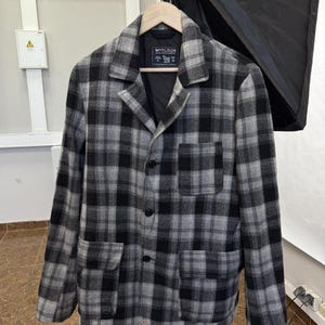 May include: A gray and black plaid jacket with a button-up front and two front pockets. The jacket is hanging on a wooden hanger. The brand name "WOOLRICH" is visible on a tag.