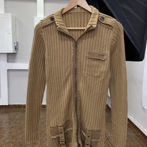 May include: A tan ribbed knit cardigan featuring a full-length front zipper, a high collar, and long sleeves. The garment includes two lower pockets with buckle straps, a single chest pocket, and shoulder epaulets with dark buttons.
