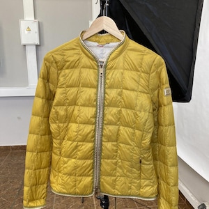 May include: A yellow quilted jacket with a gold zipper and trim. The jacket is hanging on a wooden hanger. The jacket has a stand-up collar and a small logo on the sleeve.