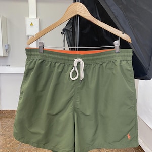 May include: Olive green swim shorts with an orange interior lining and a white drawstring. The shorts feature a small orange logo on the lower right leg. The shorts are hanging on a wooden hanger.