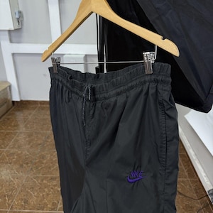 May include: Black Nike track pants hanging on a wooden hanger. The pants have an elastic waistband with a drawstring and a purple Nike logo on the left leg. The material appears to be a lightweight nylon.