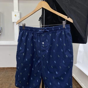 May include: Navy blue shorts with an all-over pattern of small, light blue and green polo player logos. The shorts have a drawstring waist and a single button closure. They are hanging on a wooden hanger.