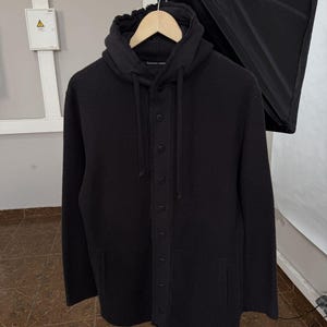 Könnte beinhalten: Eine schwarze Kapuzenjacke mit langen Ärmeln, einer durchgehenden Knopfleiste und passenden schwarzen Knöpfen. Das Kleidungsstück verfügt über eine Kapuze mit verstellbaren Kordelzügen sowie zwei Seitentaschen und besteht aus strukturiertem Material.