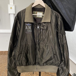 May include: Olive green bomber jacket with a light brown collar and ribbed cuffs. The jacket features a zipper closure, multiple pockets, and embroidered text and a logo on the chest. The jacket is hanging on a wooden hanger.