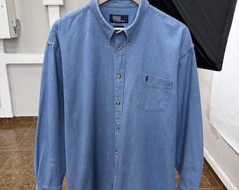 May include: A long-sleeve button-down shirt in light blue denim fabric. The shirt features a button-down collar, a single chest pocket with an embroidered dark logo, and a central row of buttons. The inner neck label reads Polo by Ralph Lauren.