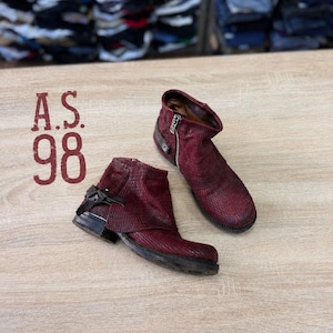 May include: A pair of dark red ankle boots with a textured finish, featuring a side zipper, a wrap-around strap with a buckle detail, and a low heel. The text A.S. 98 is printed in red on the left side of the boots.