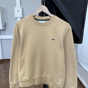 May include: A beige crewneck sweatshirt featuring a small green crocodile logo embroidered on the left chest. The garment has long sleeves, a ribbed hem, and ribbed cuffs. A white label with the brand name Lacoste is visible at the inner neck.