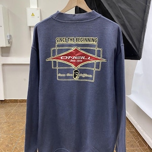May include: A navy blue long-sleeve sweatshirt featuring a graphic on the back with the text "SINCE THE BEGINNING", "O'NEILL", "Since 1952", and "Santa Cruz California". The design includes a red diamond shape, yellow outlines, and a small icon.