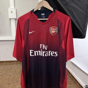 May include: A red and dark blue V-neck soccer jersey featuring the Arsenal football club crest on the left chest, a white Nike swoosh on the right, and the text Fly Emirates printed in white across the center with a wavy line pattern.