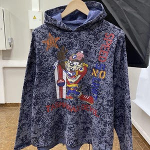 May include: A blue and dark blue acid-wash hooded sweatshirt featuring a graphic of the Tasmanian Devil character holding a snowboard. The text "SPEED ZONE" is printed vertically in red, and "TASMANIAN DEVIL" appears in red at the bottom.