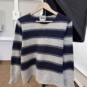 May include: A navy blue and gray striped sweater with the brand name "Acne" on the label. The sweater is made of a soft material and has long sleeves. The sweater is hanging on a wooden hanger.