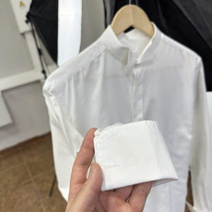 May include: A crisp white long-sleeved shirt featuring a mandarin collar and French cuffs. The cuff is held up to show a subtle, tonal embroidered logo detail on the fabric. The shirt is displayed on a wooden hanger against a neutral background.