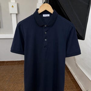 May include: A dark navy blue short-sleeved polo shirt featuring a classic collar, a three-button placket, and ribbed sleeve cuffs. The interior collar label displays the text Gran Sasso. The shirt is displayed on a wooden hanger.