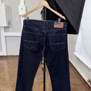 Op de afbeelding: Donkerblauwe denim jeans hangen aan een houten hanger. De jeans hebben een klassiek ontwerp met vijf zakken en een leren patch met een Amerikaanse vlag. De jeans worden gepresenteerd tegen een neutrale achtergrond.