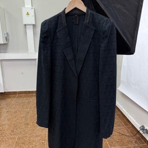 May include: A dark navy blazer with a textured pattern, hanging on a wooden hanger. The blazer has a notched lapel and a leather collar. The garment is displayed against a white background.