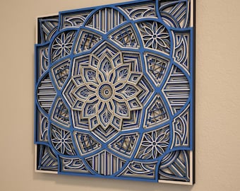 Puede incluir: Una pieza de arte mural cuadrada de madera en capas con un diseño de mandala geométrico. La obra de arte presenta intrincados patrones en tonos de azul, blanco y beige. La pieza está montada sobre un marco negro, creando un efecto dimensional.