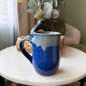 May include: A handmade ceramic mug with a deep blue glaze and a light brown rim. The mug has a curved handle and sits on a white marble table. The background includes a vase with greenery.