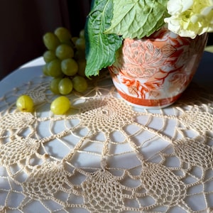 May include: A still life featuring a cream-colored, crocheted doily with a floral design. A vase with a red and orange floral pattern holds white flowers and green leaves. Green grapes are scattered around the doily.