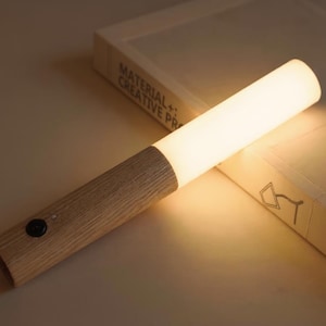 Peut inclure: Lampe LED cylindrique moderne avec manche en bois clair et lumière blanche. La lampe est posée sur une surface claire à côté d'un livre avec le texte "MATERIAL+ CREATIVE PR". La lampe mesure environ 25 cm de long.