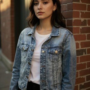 May include: A light blue denim jacket with a classic design, featuring button closures and pockets. The jacket is worn over a white t-shirt and paired with black pants. The model is standing in front of a brick wall.