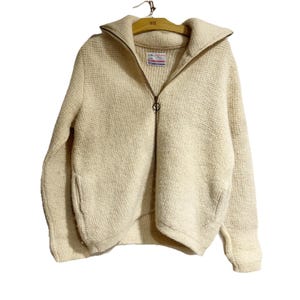 May include: Cream-colored zip-up sweater with a textured knit. The sweater features a high collar, a full-length zipper, and a small pocket on the left side. The sweater is hanging on a wooden hanger.