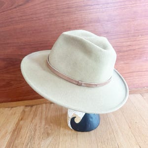 May include: A light beige fedora hat with a structured crown and a wide brim. The hat features a thin, smooth, matching beige band encircling the base of the crown, providing a clean and uniform appearance across the entire felt-like surface.