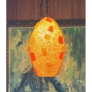 May include: A hanging pendant lamp with an oval-shaped shade featuring a textured, speckled yellow surface embedded with rectangular orange and amber glass accents. The light fixture is suspended from a dark metal chain against a dark wood wall.