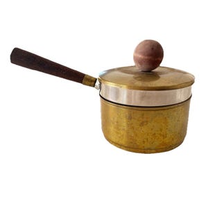 May include: A vintage brass saucepan with a wooden handle and lid. The pot has a silver band around the middle and a wooden knob on the lid. The handle is dark brown and angled.