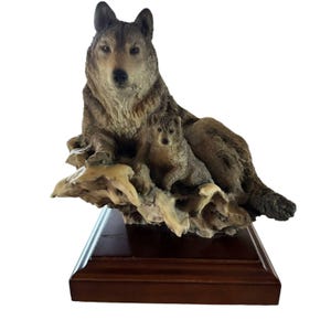 May include: A detailed figurine of a mother wolf and her cub. The wolves are a mix of brown and gray tones, resting on a cream-colored, textured base. The base is mounted on a dark brown, rectangular wooden stand. The figurine is a decorative item.