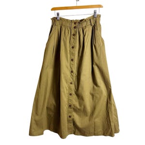 May include: A khaki-colored midi skirt featuring a high-waisted elastic waistband with belt loops, a full A-line silhouette, and a vertical row of buttons running down the center front. The skirt includes two side pockets and a gathered waistline.