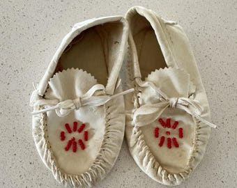 May include: A pair of off-white leather moccasins featuring a gathered toe seam, a decorative white lace tie, and a circular pattern of small red beads centered on the top of each shoe. The footwear has a soft construction and a simple slip-on design.