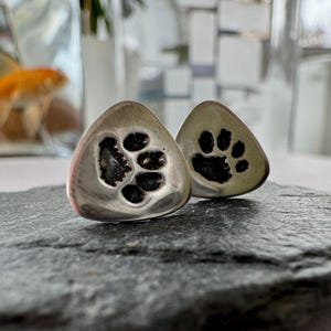 May include: Two silver triangular pendants with a paw print design. The pendants are on a dark gray surface.