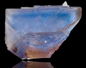 May include: A close-up of a translucent, multi-faceted mineral specimen. The crystal displays a gradient of colors, with shades of blue and hints of orange and brown. The mineral has a reflective surface, and the background is black.