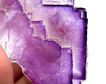 May include: A close-up of a purple fluorite mineral specimen. The crystalline structure is visible, with a mix of light and dark purple hues. The edges of the mineral are sharp and angular, with a translucent quality.