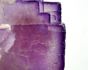 May include: Close-up of a purple fluorite crystal formation. The crystal has a layered, geometric structure with varying shades of purple and a translucent quality. The edges are rough and textured, with some white crystalline areas.
