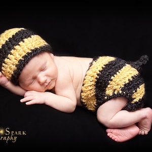 May include: A newborn baby wearing a yellow and black crocheted bee costume, sleeping on a black background.