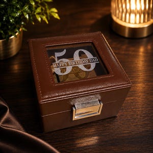 May include: A brown leather watch box with a clear top, engraved with "50 HAPPY BIRTHDAY DAD". The box has a silver clasp and sits on a dark wooden surface. A small plant and a lit candle are in the background.