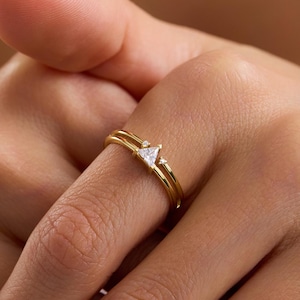 May include: A gold ring featuring a thin, polished band and a small, clear triangular gemstone set at the center. The design consists of two parallel bands that meet at the point where the triangular stone is securely mounted on the top.